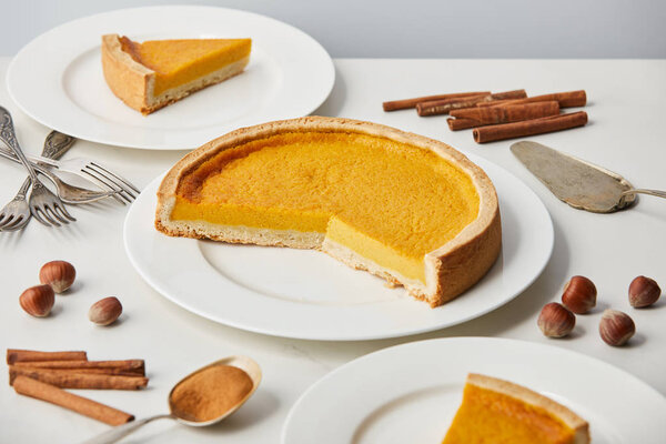 белые тарелки с вкусным тыквенным пирогом рядом со столовыми приборами и специями, изолированными на сером
