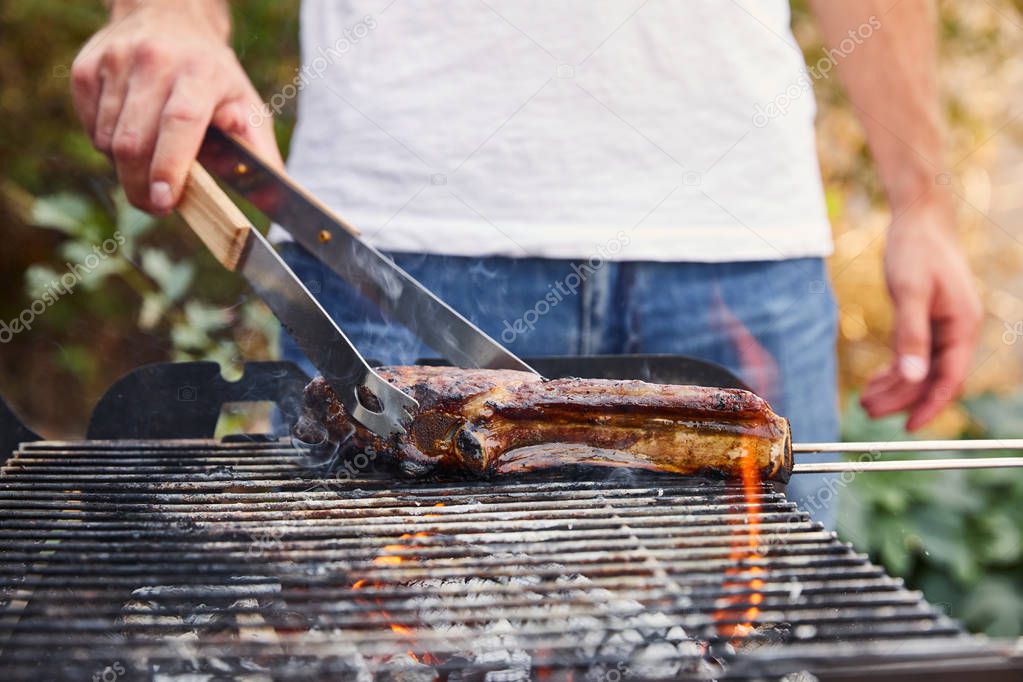 cropped view of man with tweezers grilling meat on barbecue grid 