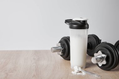Dambılların yanında protein tozu bulunan kaşık ve gri üzerine izole edilmiş spor şişesi.