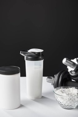 Dambıl yanında protein tozu bulunan cam kase ve siyah üzerine spor şişesi. 