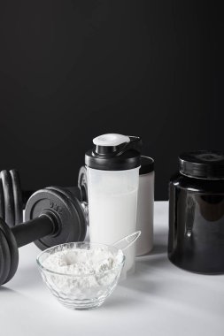 Dambıl ve spor şişesinin yanındaki cam kasede siyah protein içeceği var. 