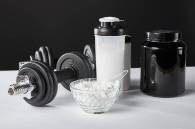 Dambıl ve spor şişesinin yanında siyah üzerine protein içeceği olan bir kase. 