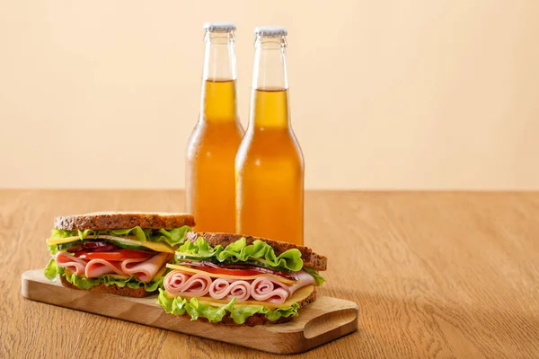 夹着生菜 熏肉和西红柿的新鲜三明治在米黄色孤立的木桌旁的啤酒瓶旁 — 图库照片