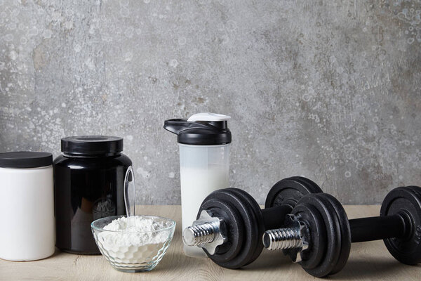 dumbbells near protein shake in sports bottle and protein powder near concrete wall 