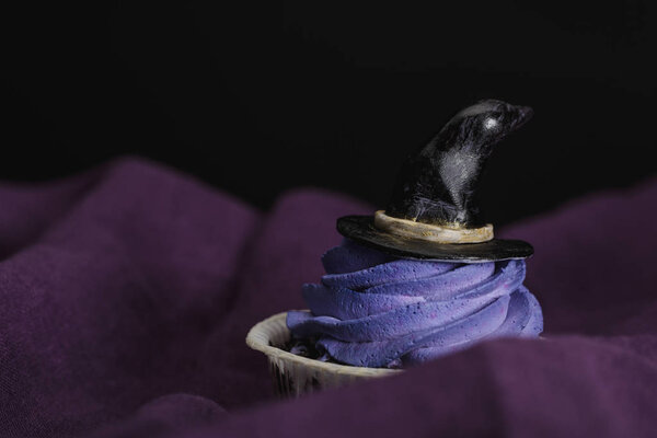 tasty Halloween cupcake with blue cream and decorative witch hat on purple cloth isolated on black