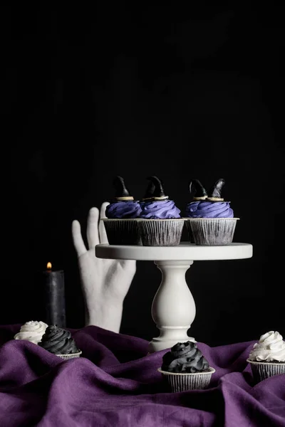 精美的万圣节纸杯蛋糕放在白色的摊子上 靠近燃烧的蜡烛 装饰的手放在紫色的布上 与黑色隔离 — 图库照片