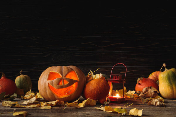 carved spooky Halloween pumpkin, autumnal leaves and burning candle on wooden rustic table on black background