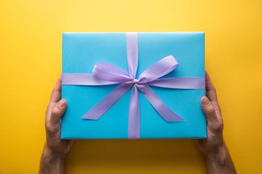 cropped view of man holding blue gift box with violet ribbon on yellow background clipart