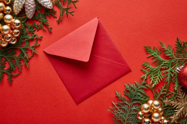 Kırmızı arkaplanda yeşil thuja dallarının parlak Noel süslemesi yakınındaki kırmızı zarfın üst görüntüsü