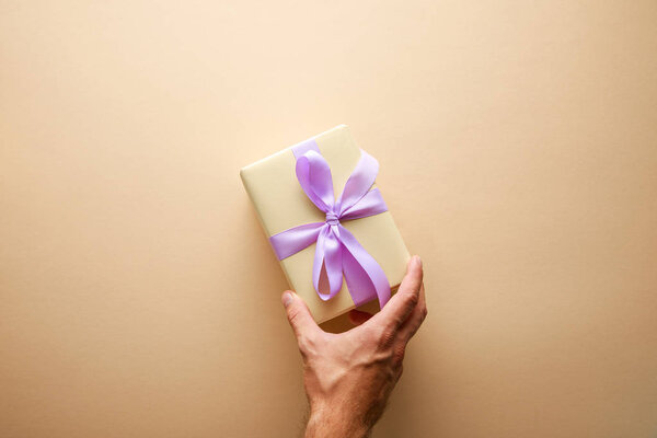 cropped view of man holding gift box with violet ribbon on beige background