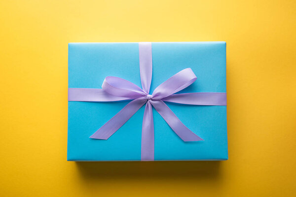 вид сверху на голубую подарочную коробку с фиолетовой лентой на желтом фоне
