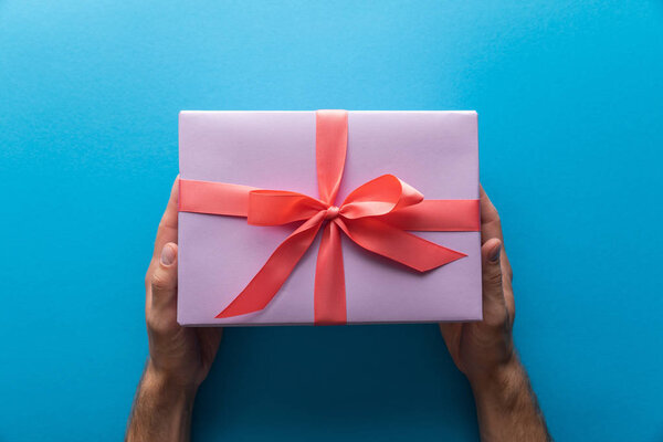 cropped view of man holding violet gift box with red ribbon on blue background