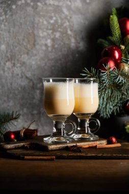 Gri taş arka planda süslü ladin dalının yanındaki kesme tahtasının üzerinde nefis eggnog içeceği olan bardaklar.