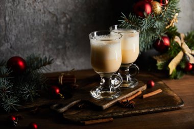 Gri taş arka planda süslü ladin dalının yanındaki kesme tahtasının üzerinde nefis eggnog içeceği olan bardaklar.