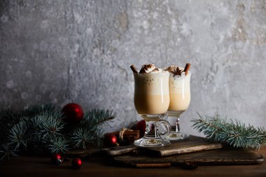 Krem şantili ve tarçınlı nefis eggnog kokteyli gri taş arka planda ladin dallarının yanındaki tahtayı kesiyor.