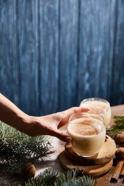 Mavi ahşap arka planda ladin dallarının yanında eggnog kokteyli ile cam içen bir kadın görüntüsü.