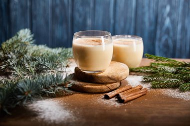 Lezzetli eggnog kokteyli ladin dalları, tarçın çubukları ve mavi desenli arka planda dağınık şeker tozu yakınlarındaki yuvarlak ahşap tahtalarda. 