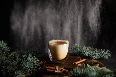 Spruce dallarının yanındaki tahtada nefis eggnog kokteyli ve siyah zemin üzerinde tarçın çubukları ve kar tanesi gibi pudra şekeri.