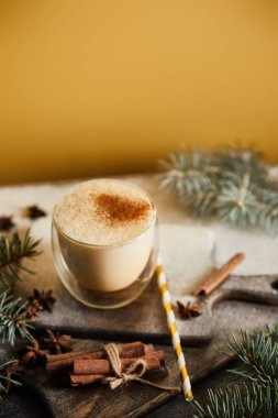 Şeker tozu, ladin dalları, tarçın çubukları ve turuncu arka planda samanla kaplı doğrama tahtasında geleneksel eggnog kokteyli.