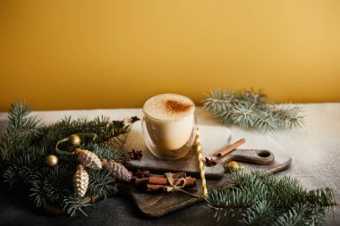 Şeker tozu, ladin dalları, tarçın çubukları, saman ve turuncu arka planda noel süslemeleri ile kaplı doğrama tahtasındaki eggnog kokteyli.
