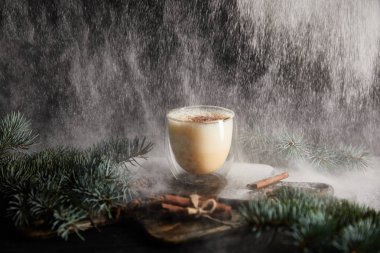 Spruce dallarının yanındaki tahtada nefis eggnog kokteyli ve siyah zemin üzerinde tarçın çubukları ve kar tanesi gibi pudra şekeri.
