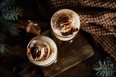 Tarçın çubukları, ladin dalları ve damalı kumaşların yanındaki tahtanın üzerinde krem şantili eggnog kokteylinin üst görüntüsü
