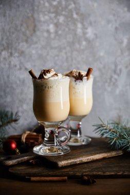 Çırpılmış krema ve tarçınlı geleneksel eggnog kokteyli gri taş arka planda ladin dallarının yanındaki tahtayı kesiyor.