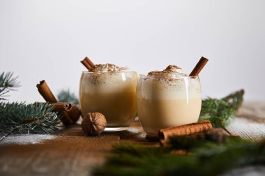 Tarçın çubuklarının yanında krem şantili eggnog kokteylinin seçici odak noktası ve ahşap masa üzerinde izole edilmiş ladin dalları.