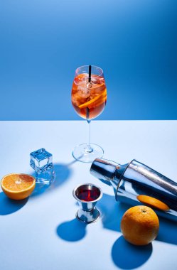 Aperol Spritz, oranges, shaker, ice cubes and measuring cup on blue background  clipart
