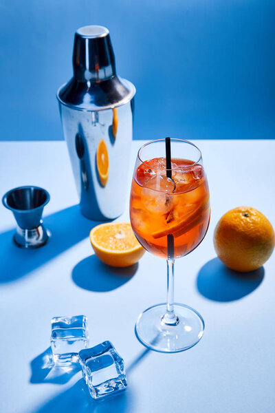 high angle view of cocktail Aperol Spritz, oranges, shaker, ice cubes and measuring cup on blue background 