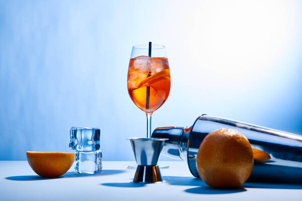 Aperol Spritz, oranges, shaker, ice cubes and measuring cup on blue background 