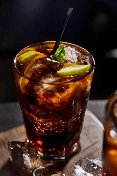 selective focus of cocktail cuba libre in glass with straw, ice cubes on black background 