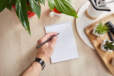partial view of man writing in notepad near green plants on wooden surface clipart
