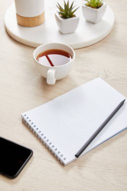 yeşil bitkiler, bir fincan çay ve ahşap yüzey üzerinde akıllı telefonun yanında kalemle boş bir defter.