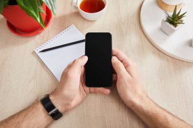 Yeşil bitkilerin yanında akıllı telefonu tutan bir adam, bir fincan çay ve tahta yüzeyde kurşun kalemle boş bir defter.