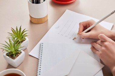 Yeşil bitkilerin yanında kağıda plan yazan kadın manzarası, bir fincan çay, zarf, bej üzerine boş bir defter.
