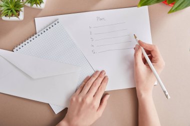 cropped view of woman writing plan on paper near green plants, envelope, blank notebook on beige surface clipart