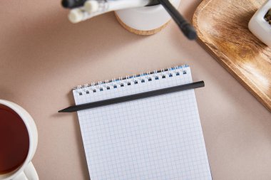 Çay fincanının üst görüntüsü, bej yüzeyinde kalemleri olan boş bir defter, seçici odaklı.