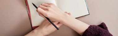 cropped view of woman writing in notebook on beige surface, panoramic shot clipart