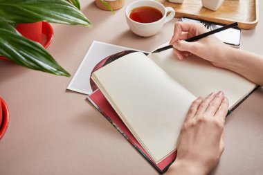 cropped view of woman writing in planner near green plant, cup of tea, smartphone on beige surface clipart