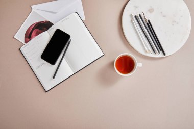 En üst çay, zarf, planlayıcı, kart, akıllı telefon, bej yüzey üzerinde kalemlerle daire içine alınmış.