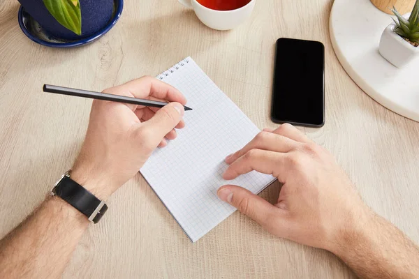 Vista Cortada Homem Escrevendo Notebook Perto Smartphone Superfície Madeira — Fotografia de Stock