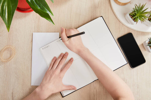 cropped view of woman writing in notebook near green plants and smartphone on wooden surface