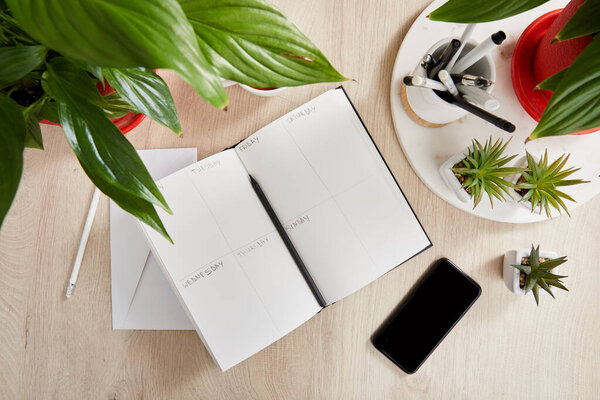 top view of green plants, blank notebook with pencils and pens near smartphone on wooden surface