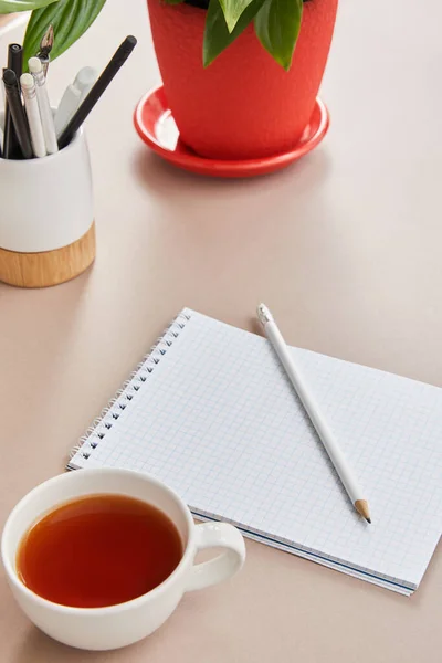 green plant, cup of tea, blank notebook with pencils on beige surface