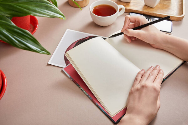 обрезанный вид женщины, пишущей в планировщике возле зеленого растения, чашка чая, смартфон на бежевой поверхности
