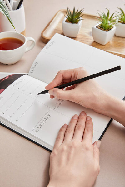 обрезанный вид женщины, пишущей в планировщике рядом с зелеными растениями на деревянной доске, чашка чая на бежевой поверхности

