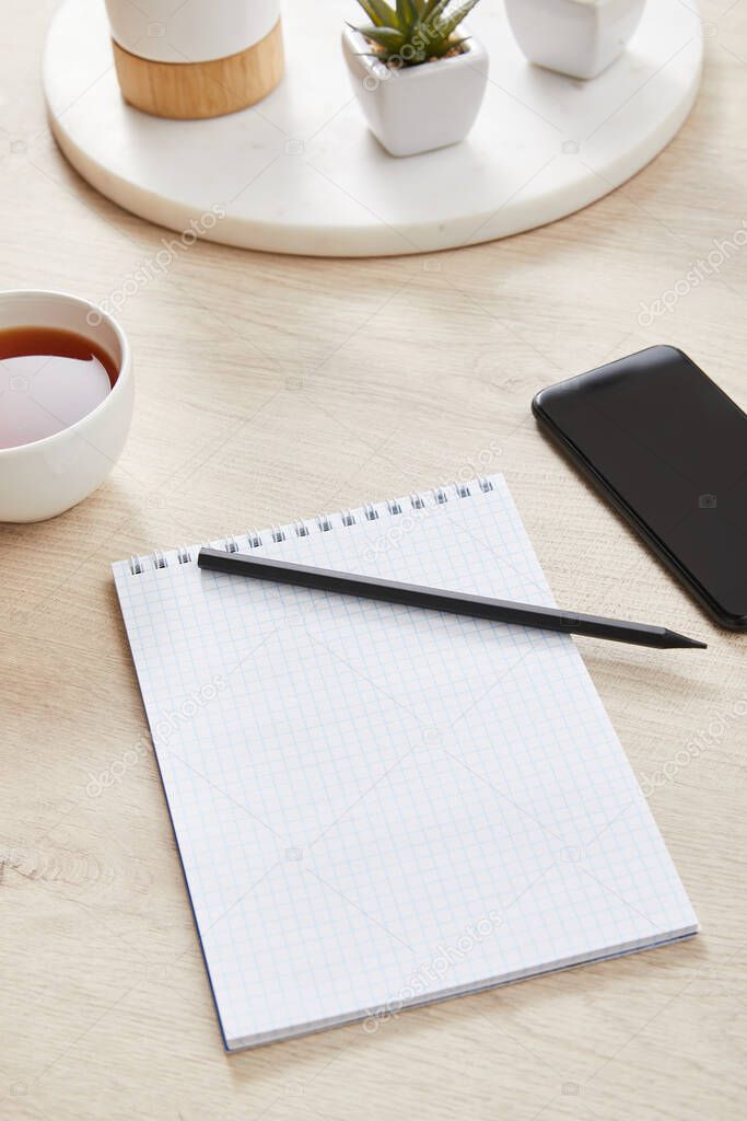 green plant, cup of tea and blank notebook with pencil near smartphone on wooden surface