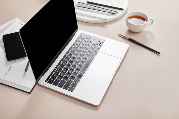 お茶と文房具のカップの近くに空白の画面とベージュの表面にスマートフォンを持つノートパソコン — ストック写真