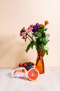 floral and fruit composition with bouquet in vase, berries, grapefruit and apricots on wooden surface on beige background clipart